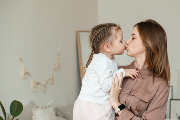 A little girl kisses her mother at home in a modern bright bedroom. The concept of parenthood, love...