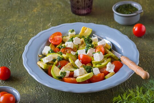 Summer salad with grilled zucchini, tomatoes, feta cheese and parsley and dill pesto in a gray plate on a green concrete background. Salad recipes.