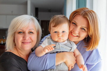 Ukrainian mother with infant daughter and grandmother stands near window in home