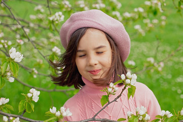 A happy little girl stands with her eyes closed in a blooming white apple tree in the park.