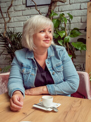 Senior woman in denim jacket sits at table and drink coffee in cafe