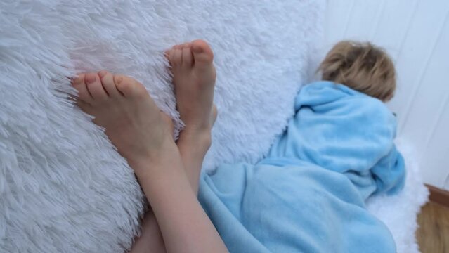 The teenage girl sat comfortably on the couch. The child is dressed in a blue house robe. In the foreground are bare children's feet and a fluffy blanket. The child plays in the smartphone. Social