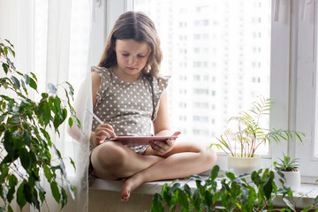 Cute little girl sitting near window at home and drawing with a digital tablet and a pen stylus.