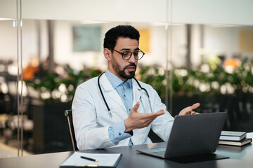 Serious millennial islamic man doctor with beard in glasses, white coat speaks with patient at laptop in clinic
