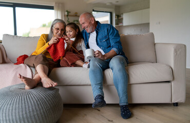 Obraz premium Little girl sitting on sofa with her grandparents and learning to knit indoors at home.