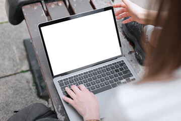 A laptop with a blank white screen stands on a bench. A girl types something on a laptop while sitting on a bench outside. Mockup laptop.