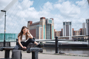 Fototapeta na wymiar A young girl works at a laptop sitting on a bench, against the backdrop of the Gateshead Millennium Bridge. The freelancer works outdoors. The concept of a free office. North East England