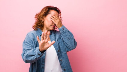 middle age hispanic woman covering face with hand and putting other hand up front to stop camera,...