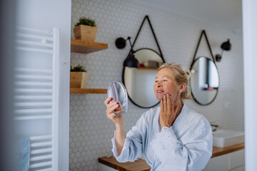 Beautiful senior woman in bathrobe looking at mirror and applying natural face cream in bathroom,...