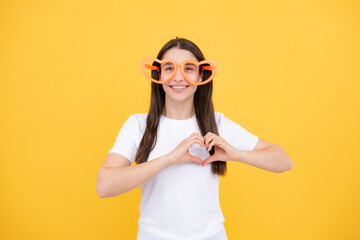 Hearts style. Portrait of a funny girl wearing cool party glasses. Cheerful young girl smiling with heart-shape glasses on yellow background.