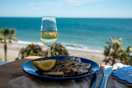 Sardines espeto prepared on skewers and open flame on fireplace with olive trees wood, served outdoor with glass of fino sherry and view on blue sea, Malaga, Andalusia, Spain
