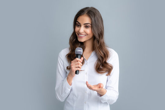 Beautiful Business Woman Is Speaking On Conference. Happy Smiling Business Woman Holding Mic, Standing With Microphone Against Grey Background, Wearing Shirt.