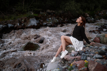 Cute brunette woman sits on the stones next to the river. in nature. Portrait of an elegant woman in black and white clothes in nature as advertising cover or promotion design.