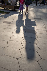 The feet of strolling pedestrians on the sidewalk paved with hexagonal large tiles. Human shadow on...
