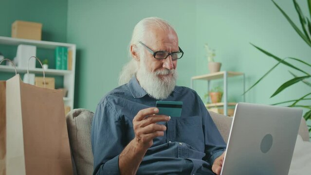 Overjoyed Elderly Man Sitting at the Sofa with a Laptop Holding Credit Card and Making a Purchase from the Internet. Older Grandparent Learn Using Modern Technology Gadget Concept.