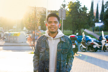 waist up shot of a originarian mexican feature man standing with hands in pockets on a urban place with land vehicles and the sun lights behind him