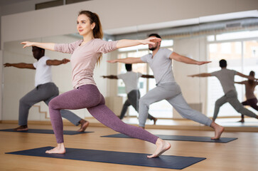 Fototapeta premium Concentrated young woman standing in Revolved High Lunge Pose during yoga class with group in fitness studio..