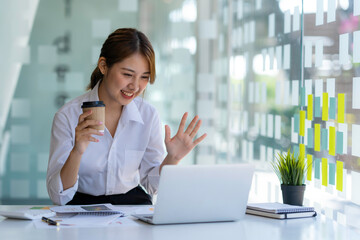 Happy young asian business woman waving hands to greeting partner during making video conference with her team.