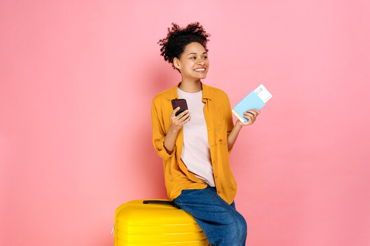 Joyful african american curly haired girl, holds passport with travel tickets and smart phone, sit on yellow suitcase on isolated pink background, looking to the side, dreams about vacation, smiling