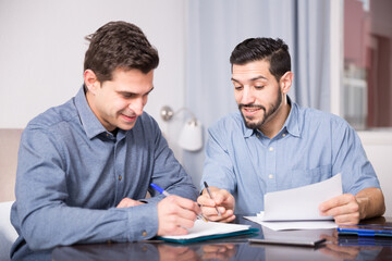 Portrait of two cheerful young men reading documents together at home table - Powered by Adobe