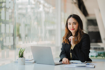 Fototapeta premium Smiling beautiful Asian businesswoman sitting with laptop and computer working on paperwork make an account analysis report real estate investment information financial and tax system concepts