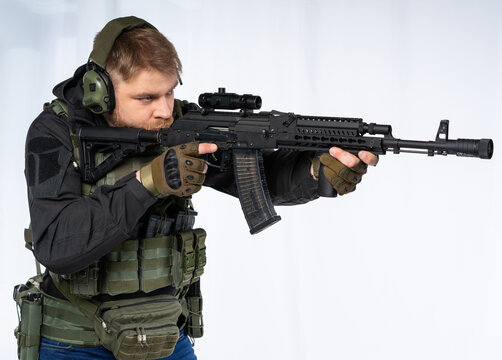 airsoft player in full gear with fire series guns. a man in headphones, a bulletproof vest, with a backpack and a belt, aims his machine gun to the side. profile. White background.