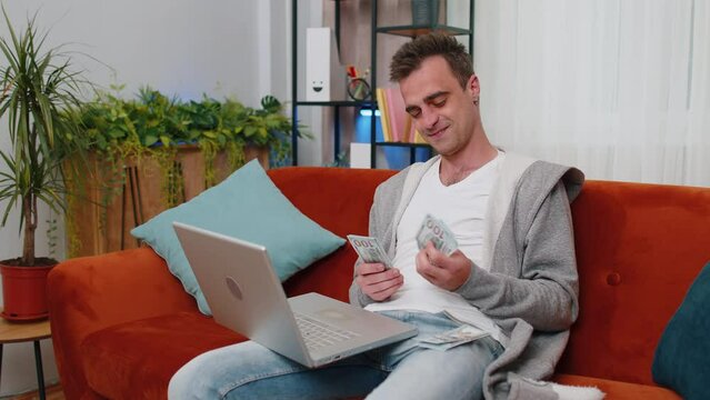 Planning budget. Smiling rich happy young man counting money cash use laptop computer calculate domestic bills at home. Joyful guy satisfied of income earnings, saves money for planned vacation, gifts