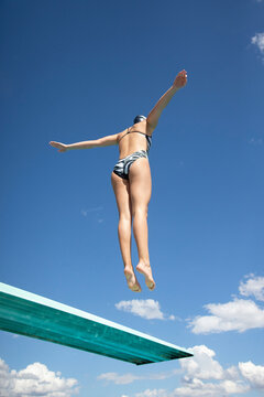 View from below. Fit woman jumping high off a diving board into the swimming pool. Abstract image with blue sky and clouds in the background. Lots of copy space.