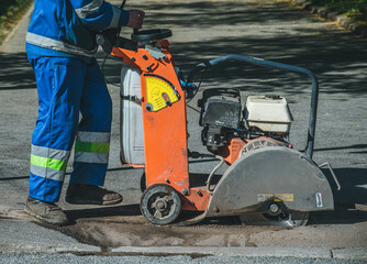 A road worker in overalls uses a portable asphalt cutter to cut asphalt with a diamond blade to...