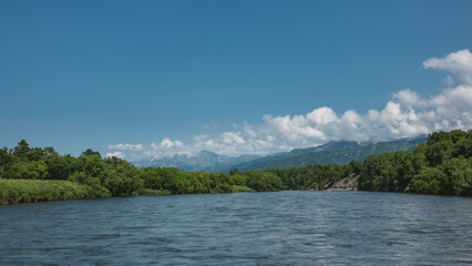 Fototapeta premium A calm blue river flows under an azure sky. A rafting boat is visible in the distance. Lush green vegetation on the banks. A mountain range against the background of clouds. Kamchatka
