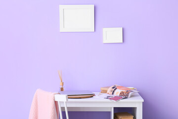 Workplace with laptop, magazine and blank frames on lilac wall