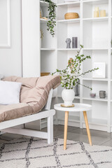 Vase with blooming branches on table, couch and shelving unit in light living room
