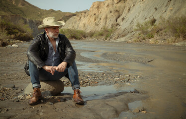 Adult man in cowboy hat sitting on rock along river flows in desert. Almeria, Spain