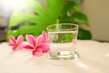 Obraz na płótnie Canvas Glass Mineral Water On Sand Next To Pink Frangipani Flowers.