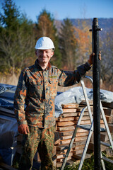 Naklejka premium Male worker building pile foundation for wooden frame house. Portrait of man builder in white safety helmet drilling piles into the ground on sunny day.