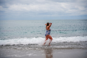 Fototapeta premium A plump woman in a bathing suit enters the water during the surf. Alone on the beach, Gray sky in the clouds, swimming in winter.