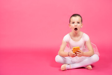 Surprised little ballet dancer girl holding phone and looking at camera isolated on pink background.