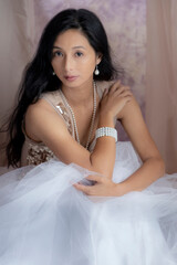 A beautiful Asian woman wearing a gold sequin top and tulle skirt with pearl jewellery and surrounded by draped fabric