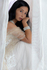 Beautiful Asian woman with long hair wearing a white lace dress and pearl jewellery surrounded by...