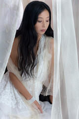 Beautiful Asian woman with long hair wearing a white lace dress and pearl jewellery surrounded by...