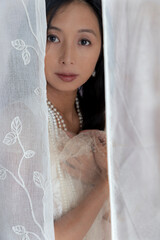 Beautiful Asian woman with long hair wearing a white lace dress and pearl jewellery surrounded by...
