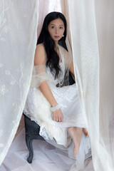 Beautiful Asian woman with long hair wearing a white lace dress and pearl jewellery surrounded by...