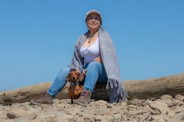 Mature female hiker sitting on a log taking a break next to her brown dachshund, Maasvallei nature reserve, blue sky in the background, sunny day in South Limburg, Netherlands. Active life concept