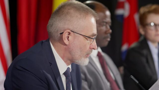 Tilt up shot of senior Caucasian diplomat sitting at press conference, reading statement from paper and answering questions of journalists