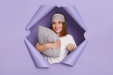 Horizontal shot of smiling delighted woman breaks through purple paper background, holding pillow,...