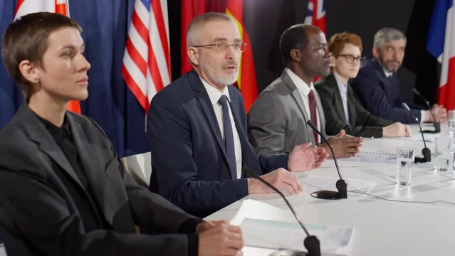 Senior Caucasian politician sitting at press conference table with multiethnic participants and speaking in mic while answering questions of journalists