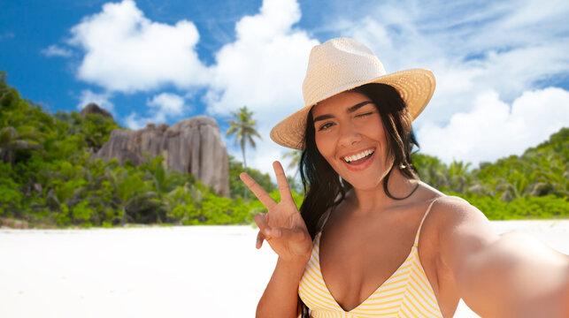 people, summer and swimwear concept - happy smiling young woman in bikini swimsuit and straw hat taking selfie and showing peace gesture over seychelles island beach background