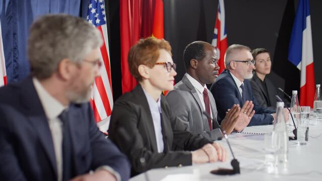 Rack focus shot of multiethnic male and female diplomats sitting at press conference table and speaking in mics in front of journalists