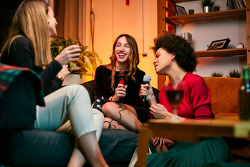 A multicultural group of female friends sings karaoke while drinking alcohol in the living room.
