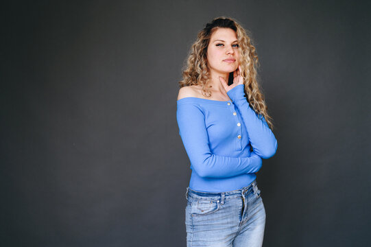 Horizontal photo of a curly blonde woman in a blue sweater, jeans, standing sideways, arms folded on her chest